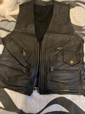 Svart skinnväst med dragkedjor - Säljer min pappas svart väst i skinnimitation med flera dragkedjeförsedda fickor och en ficka med tryckknapp fram. Västen har en rak passform och stängs med dragkedja. Snygg detalj med snörning i sidorna. Perfekt för dig som gillar en tuffare stil. 100% skinn
