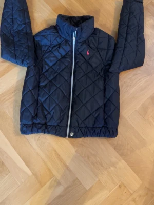 Mörk blå quiltad jacka från Polo Ralph Lauren - Snygg blå quiltad jacka från Polo Ralph Lauren med dragkedja framtill och klassisk röd logga på bröstet. Jackan har hög krage och elastisk nederkant för en bekväm passform. Perfekt för kyligare dagar.