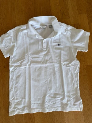 Vit piké t-shirt från GANT, 158/164 - Klassisk vit piké t-shirt från GANT med korta ärmar och liten broderad logga på bröstet. Tröjan har krage och knappar vid halsen. Perfekt för en clean och stilren look. Storlek 158/164