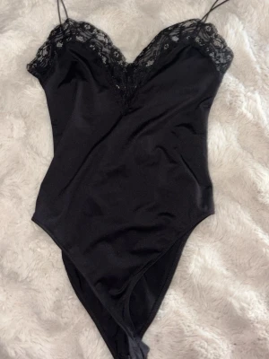Svart spetsbody från Zara - Snygg svart body från Zara med tunna axelband och v-ringning dekorerad med spets. Tillverkad i mjukt, stretchigt material som sitter skönt mot kroppen. Perfekt för att skapa en trendig och elegant look.
