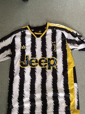 Juventus matchtröja svart/vit/gul - Juventus matchtröja från Adidas med svartvita borstade ränder och gula detaljer. Jeep-logga på bröstet, klubbmärke med två stjärnor och Adidas-logga. Tillverkad i lätt och ventilerande material, perfekt för fotboll eller träning.