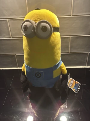 Minion mjukisdjur gul och blå - Roligt Minion mjukisdjur köpt på elgiganten för 299 kr. i gult och blått med stora grå glasögon och svarta handskar. Figuren har blå overall med Gru-logga på magen och en etikett med Minion-motiv. Perfekt för dig som gillar Despicable Me!