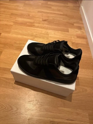 Svarta maison margiela gats - Säljer ett par stilrena svarta sneakers i en kombination av mocka och slätt läder. Skorna har klassisk låg profil, svarta skosnören och en diskret design som passar till det mesta. Perfekta för dig som gillar minimalistisk och clean stil.