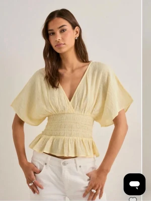 Ljusgul omlottopp med volang - Superfin ljusgul topp med omlott och v-ringning. Vida ärmar, smockad midja och volang nertill ger en söt och trendig look. Perfekt till jeans eller kjol för en fräsch stil.