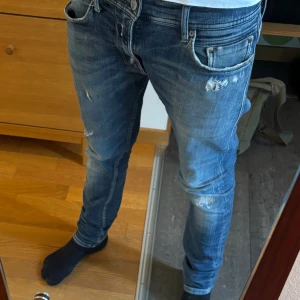 Replay Jondrill blå jeans med slitningar - Säljer ett par Replay Jondrill jeans i blå tvätt med snygga slitningar och lätt distressed look. Jeansen har klassisk femficksdesign, smal passform och läderpatch bak i midjan. Perfekta för dig som gillar en modern och avslappnad stil. Storlek W32 L32 