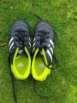 Svarta Adidas fotbollsskor med neon - Säljer ett par klassiska Adidas fotbollsskor i svart med vita ränder och neonfärgade detaljer. Skorna har snörning, tålig ovandel i syntetmaterial och dobbar för spel på gräs. Insidan är fodrad i neonfärg för extra stilpoäng. Pris kan diskuteras 
