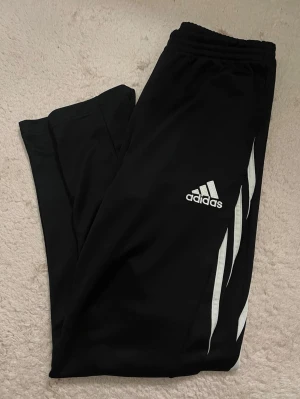 Svarta träningsbyxor från Adidas - Svarta träningsbyxor från Adidas med vita klassiska ränder längs benen och Adidas-logga på låret. Byxorna har resår i midjan och är tillverkade i ett lätt och bekvämt material, perfekta för träning eller chill. Använda fåtal gånger, som ny🩷