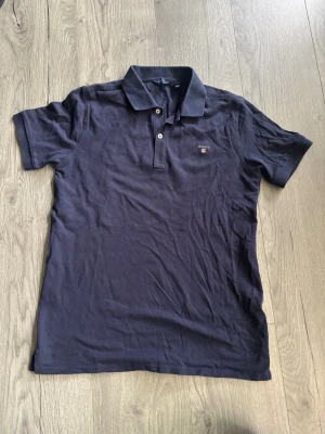 Marinblå pikéskjorta från Gant - Tja! Jag säljer en marinblå piké t-shirt från Gant med klassisk krage och två knappar framtill. Tillverkad i mjuk bomull och har korta ärmar samt en liten broderad logga på bröstet. Enkel och stilren modell. Pikén är i fint även fast den använts ett tag! Inget fel på den osv.