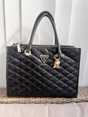 Svart quiltad handväska från Guess - Snygg svart handväska från Guess med quiltat mönster och guldfärgade detaljer. Väskan har dubbla handtag, dragkedja upptill, samt en praktisk ytterficka med dragkedja på baksidan. Guess-logga framtill och dekorativa nyckelringar. Tillverkad i konstläder. Den är nästan helt oanvänd!