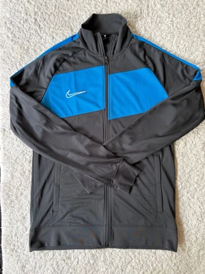 Nike träningsjacka blå och grå - Nike träningsjacka i grått och blått med vit broderad Swoosh-logga på bröstet. Jackan har hel dragkedja, ståkrage och två sidofickor. Tillverkad i ett lätt och funktionellt material som passar perfekt till träning eller uppvärmning.
