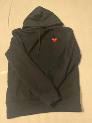 Svart cdg hoodie  - Säljer en svart hoodie med huva och dragsnören. På bröstet finns en broderad patch med ett rött hjärta och ögon, vilket ger en unik och trendig look. Hoodien har magficka och är tillverkad i mjukt bomullstyg. Perfekt för dig som gillar streetwear och vill sticka ut.