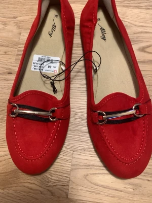 Röda loafers med metallspänne - Snygga röda loafers från Alley i storlek 38. Skorna har en klassisk form med rundad tå och dekorativt metallspänne framtill. Tillverkade i mockaimitation med kontrastsömmar och diskret svart banddetalj. Perfekta för en trendig och avslappnad look.