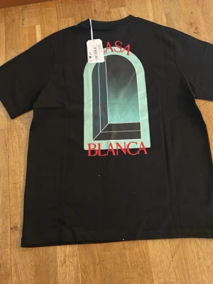 Svart Casa Blanca t-shirt med tryck - Cool svart t-shirt med stort grafiskt tryck i turkos och rött på ryggen samt mindre tryck på bröstet. Rund hals och korta ärmar. Perfekt för dig som gillar streetwear och vill sticka ut lite extra.