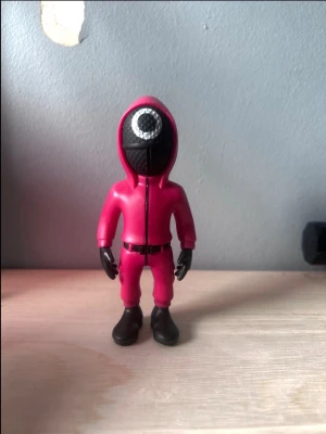 Squid Game maskeradmask med huva - Maskeradmask inspirerad av Squid Game med svart ansikte och vit cirkel, fastsydd på en röd huva och overall. Tillverkad i plast och tyg, perfekt för cosplay eller temafest. Masken täcker hela ansiktet och har detaljerade svarta handskar och stövlar.