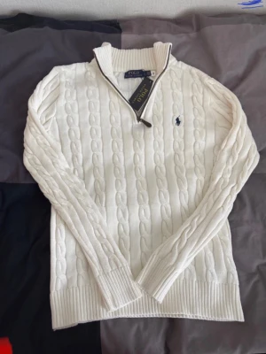 Vit kabelstickad tröja Polo Ralph Lauren - Ny oanvänd Kabelstickad vit tröja från Polo Ralph Lauren med halv dragkedja och ribbade muddar. Tröjan har den klassiska marinblå loggan broderad på bröstet och är tillverkad i mjukt material. Perfekt för en stilren och avslappnad look.ett till ett ,fraktar snabbt