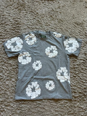 Grå t-shirt med vita blommor - Snygg grå t-shirt med stora vita blommiga tryck över hela plagget. Klassisk passform och rund halsringning. Perfekt för dig som vill sticka ut med ett unikt mönster. Tillverkad i mjuk bomull för skön känsla.