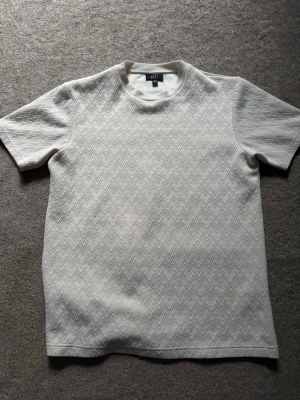 Vit t-shirt med zigzagmönster - Snygg vit t-shirt med diskret zigzagmönster över hela plagget. T-shirten har rund halsringning och korta ärmar. Perfekt för dig som gillar stilrena detaljer och en modern look.