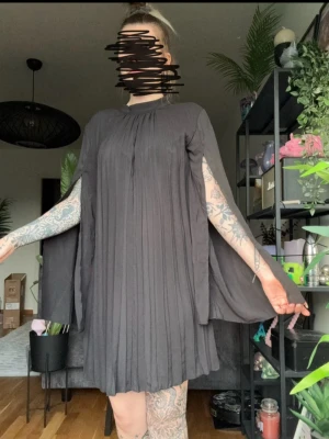 Svart Elegant Klänning  - Säljer en svart kortklänning med plisserat tyg och cape-liknande ärmar. Klänningen har rund halsringning och ett flowy fall som ger en cool och unik look. Perfekt för dig som vill sticka ut lite extra.