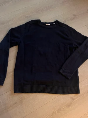 Mörkblå stickad tröja XL - Mörkblå stickad tröja från Knowledge Cotton Apparel i storlek XL. Tröjan har rund halsringning och ribbade muddar vid ärmslut och nederkant. Tillverkad i mjukt material som passar perfekt för kyligare dagar.