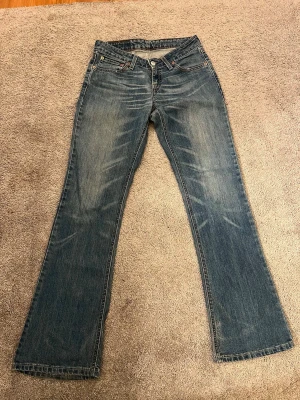 Blå bootcut jeans med slitningar - Ett par klassiska blå bootcut jeans med snygga slitningar framtill och bakfickor med dekorativa sömmar. Jeansen har fem fickor och stängs med knapp och dragkedja. Perfekta för en avslappnad och trendig look.