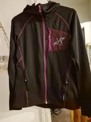 Revolution Race illusion hoodie - Snygg svart hoodie med dragkedja och lila kontrastsömmar. Har en huva och en bröstficka med dragkedja och logga på bröstet. Tillverkad i mjukt material, perfekt för chill eller träning. Lila detaljer ger en sportig vibe. Storlek xs men ganska stor i storleken. Passar s även liten m
