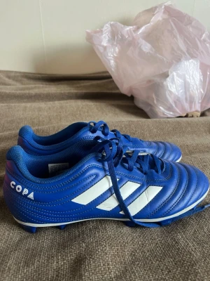Blå Adidas Copa fotbollsskor - Snygga blå Adidas Copa fotbollsskor med klassiska vita ränder på sidan och snörning. Skorna har låg profil och dobbar för spel på gräs. Tillverkade i slitstarkt syntetmaterial som ger bra komfort och kontroll på planen.