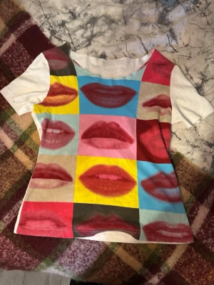 T shirt me tryck  - Unik t-shirt med korta ärmar och popkonst-inspirerat tryck av läppar i olika färger på framsidan. Basen är vit och materialet känns som bomull. Perfekt för dig som vill sticka ut med en färgstark och kreativ stil. Avklippt krage 