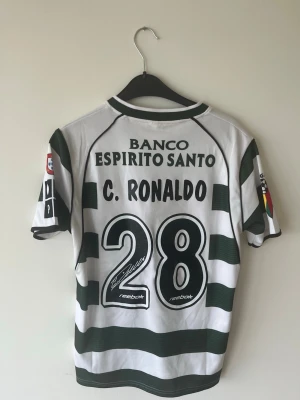 Sporting lisbon #28 cristiano ronaldo - Klassisk vit och grönrandig fotbollströja från Sporting Lissabon med C. Ronaldo och nummer 28 på ryggen. Tröjan har klubbmärke på bröstet, sponsorloggor och korta ärmar. Perfekt för dig som älskar fotboll och samlar på retrotröjor. Endast testad 