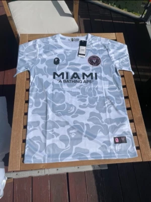 A Bathing Ape x Inter Miami t-shirt - Snygg t-shirt från A Bathing Ape i samarbete med Inter Miami. Tröjan har ett grått och vitt camouflage-mönster, rund hals och korta ärmar. Stor MIAMI-logga framtill och klassiska BAPE-apan på bröstet. Perfekt för dig som gillar streetwear och fotboll.