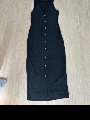Svart långklänning med knappar - Svart långklänning med v-ringning och dekorativa knappar framtill. Klänningen är ärmlös och har ett diskret hålmönster i tyget som ger en snygg struktur. Perfekt för dig som gillar enkel men stilren design.