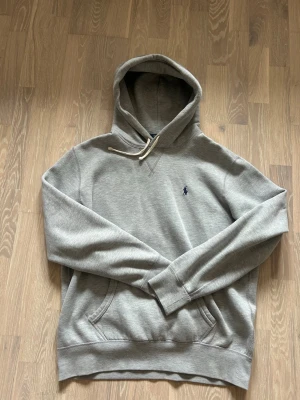 Grå hoodie från Polo Ralph Lauren - Snygg grå hoodie från Polo Ralph Lauren med klassisk huva och vita dragsnören. Broderad blå logga på bröstet och känguruficka framtill. Tillverkad i mjuk bomullsblandning som känns skön mot huden. Perfekt för en avslappnad och stilren look. Inga fläckar eller märken.