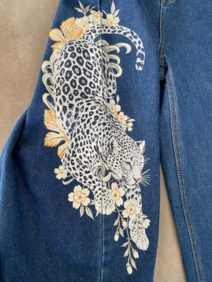 Blå baggy jeans med tryck  - Blå baggy jeans som är helt nya och aldrig använda. Dom har ett stort tryck med blommönster och en leopard på ena benet och en stor blomma på andra benet. Byxorna har hög midja och raka ben. 