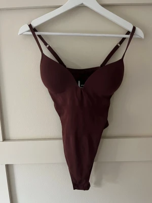 Vinröd body med tunna axelband - Snygg vinröd body med vadderade kupor och tunna, justerbara axelband. Djup urringning framtill och slät, stretchig material som formar sig efter kroppen. Perfekt att styla under skjorta eller kavaj.