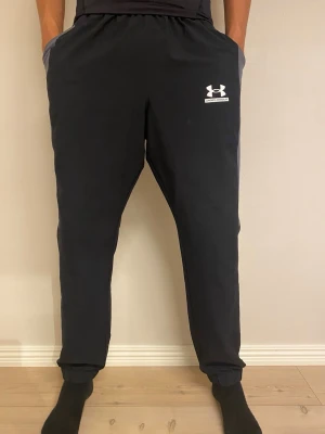 Svarta mjukisbyxor från Under Armour - Snygga svarta mjukisbyxor från Under Armour med vit logga på vänster lår. Byxorna har elastisk midja, sidofickor och smal passform med muddar vid bensluten. Perfekta för chill eller träning.