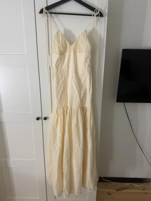 Ljusbeige klänning - Säljer en ljus, lång klänning med smala axelband och v-ringad hals. Klänningen har en volang nedtill och är tillverkad i ett tunt, mjukt tyg som känns perfekt för varma dagar. Enkel och stilren design som ger en romantisk vibe. Helt slutsåld överallt.