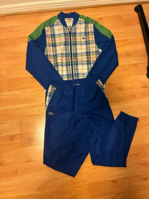 Blå och grön rutig jumpsuit Lacoste - Snygg jumpsuit från Lacoste i blått och grönt med rutigt mönster på överdelen. Dragkedja framtill, långa ärmar och ben samt elastisk midja. Materialet känns lätt och sportigt, perfekt för en chill och trendig look. Klassisk Lacoste-logga på bröstet och byxan.