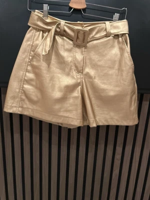 Guldiga shorts med bälte och knyt - Snygga guldiga shorts i syntetmaterial med hög midja och tillhörande bälte med knyt. Shortsen har en loose passform och sidofickor, perfekta för att sticka ut i mängden.