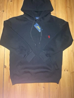 Svart hoodie från Polo Ralph Lauren - Säljer en svart hoodie från Polo Ralph Lauren med klassisk röd logga broderad på bröstet. Hoodien har huva med dragsko och en stor magficka framtill. Tillverkad i mjukt bomullsmaterial, perfekt för en avslappnad och stilren look.