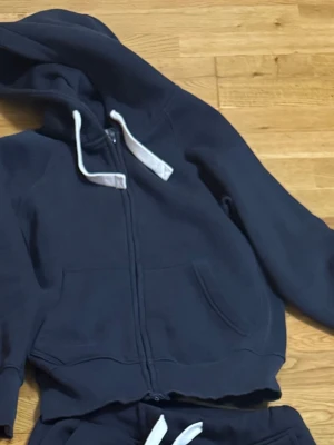 Mörkblå Russemerch hoodie med dragkedja  - En mörkblå Russemerch hoodie med dragkedja och stora fickor framtill. Utrustad med huva och vita dragsnören. 