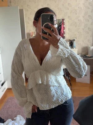 Vit strukturerad blus med volang från GinaTricot  - Supersnygg vit blus med strukturmönster och volang i midjan. Djup v-ringning och långa ärmar ger en trendig look. Perfekt att styla med jeans eller kjol för en chic outfit. Köpt för 399kr.
