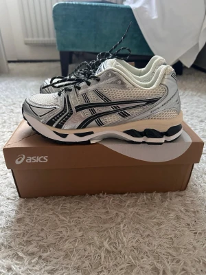 Asics vita och silver sneakers - Snygga Asics sneakers i vitt och silver med svarta detaljer. Skorna har en sportig mesh- och syntetöverdel, klassisk Asics-logga på sidan och tjock, stötdämpande sula. Perfekta för dig som gillar retrostil och komfort. Svarta och vita skosnören ingår.