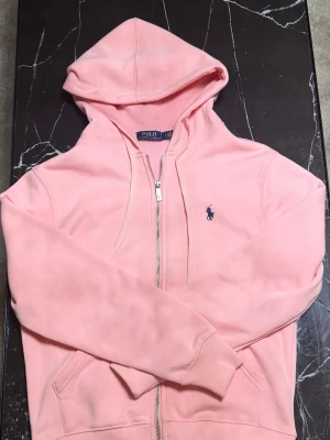 Ljusrosa hoodie från Polo Ralph Lauren - Snygg ljusrosa hoodie med dragkedja från Polo Ralph Lauren. Klassisk huva, två fickor framtill och den ikoniska blå loggan broderad på bröstet. Tillverkad i mjukt bomullsmaterial som känns skönt mot huden. Perfekt för en chill och avslappnad stil.