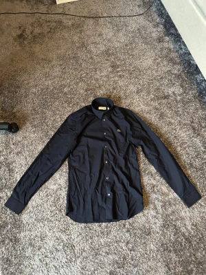 Mörkblå Lacoste Skjorta  - Sprillans ny Lacoste skjorta som aldrig är använd. Kostar 1195kr ny. Storleken är 37 och den passar xs-S och passformen är slim.