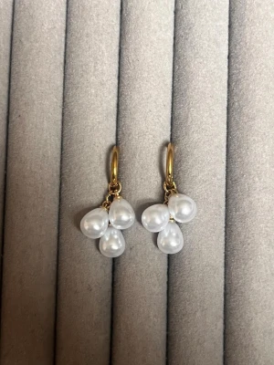 Guldörhängen med droppformade pärlor - Säljer ett par snygga örhängen med guldfärgade ringar och tre droppformade vita pärlor på varje örhänge. Perfekt accessoar för dig som gillar en elegant och trendig look. Materialet är metall och pärlorna är i plast eller glas.