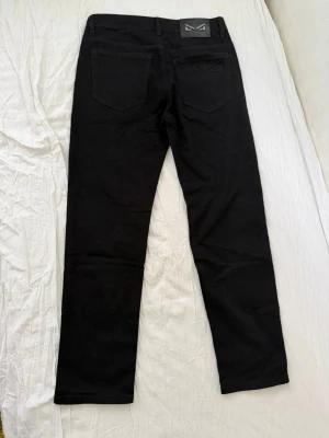 Svarta jeans med broderad fickdetalj - Snygga svarta jeans med raka ben och cool broderad detalj på ena bakfickan. Jeansen har klassisk femficksdesign och bälteshällor. Materialet ser ut att vara bomull med lite stretch för extra komfort.