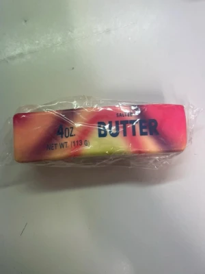 Färgglad butter squishy  - En populär butter squishy  på 113 gram med färgglada mönster i rött, gult och orange. Tvålen är inslagen i plast och har texten 'BUTTER' tryckt på sidan, vilket ger den ett lekfullt utseende. Perfekt som dekoration eller till handtvätt.