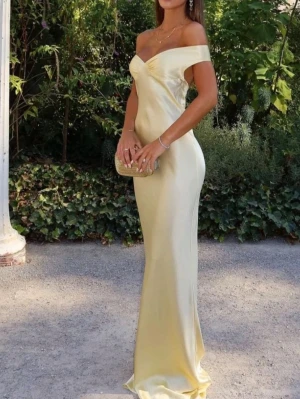 Ljusgul axelbandslös långklänning - Elegant långklänning i ljusgul satin med offshoulder-design och figurnära passform. Klänningen har en vacker v-ringning och slits i sidan för extra rörelsefrihet. Perfekt för sommarens speciella tillfällen.