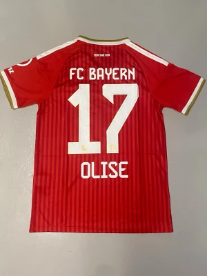 Olise#17 FC Bayern München 26/27 hemmaställ  - Säljer en röd FC Bayern München matchtröja från Adidas med vita detaljer och guldkanter. Tröjan har nummer 17 och namnet Olise på ryggen, samt klubbmärke och sponsorloggor på framsidan. 