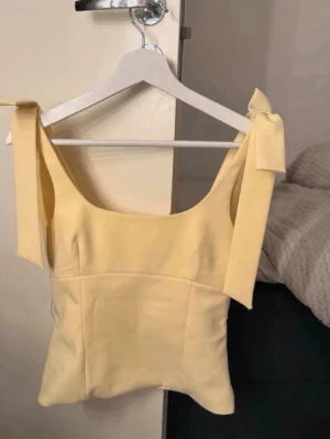 Gul topp med rosetter  - Snygg ljusgul linnetopp med breda axelband som knyts till rosetter. Toppen har en rak passform och ärmlös design, perfekt för varma dagar. Materialet känns mjukt och lätt mot huden och färgen ger en fräsch vibe till din outfit.