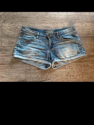 Blå denimshorts med fickor - Snygga blå jeansshorts i klassisk femficksmodell med slitna detaljer och uppvikta benslut. Perfekta för varma dagar och enkla att matcha med allt. Stängs med knapp och dragkedja framtill.